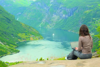 A person with a laptop, back to the camera, sits on a rock overlooking a tranquil mountain lake.